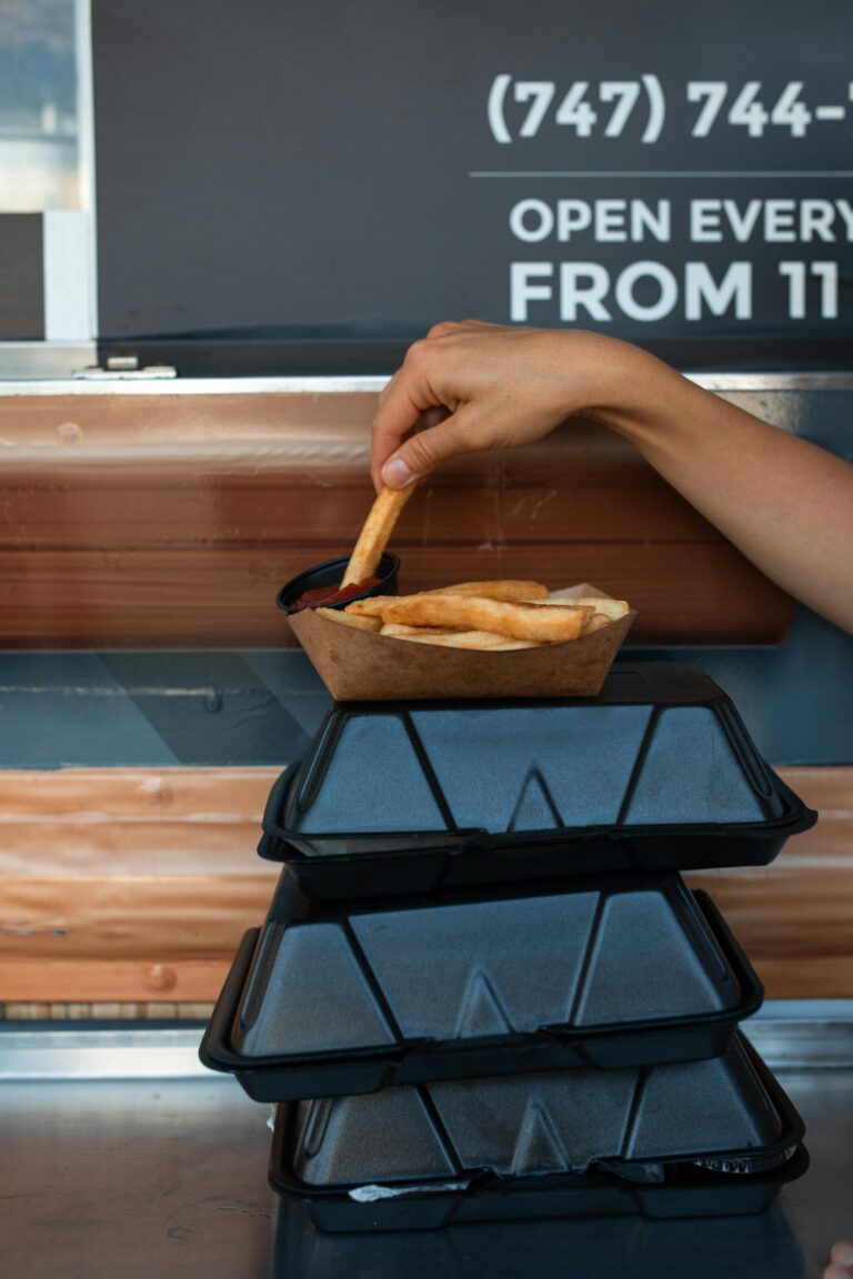 A hand holding a French fry above sauce at a food truck window.